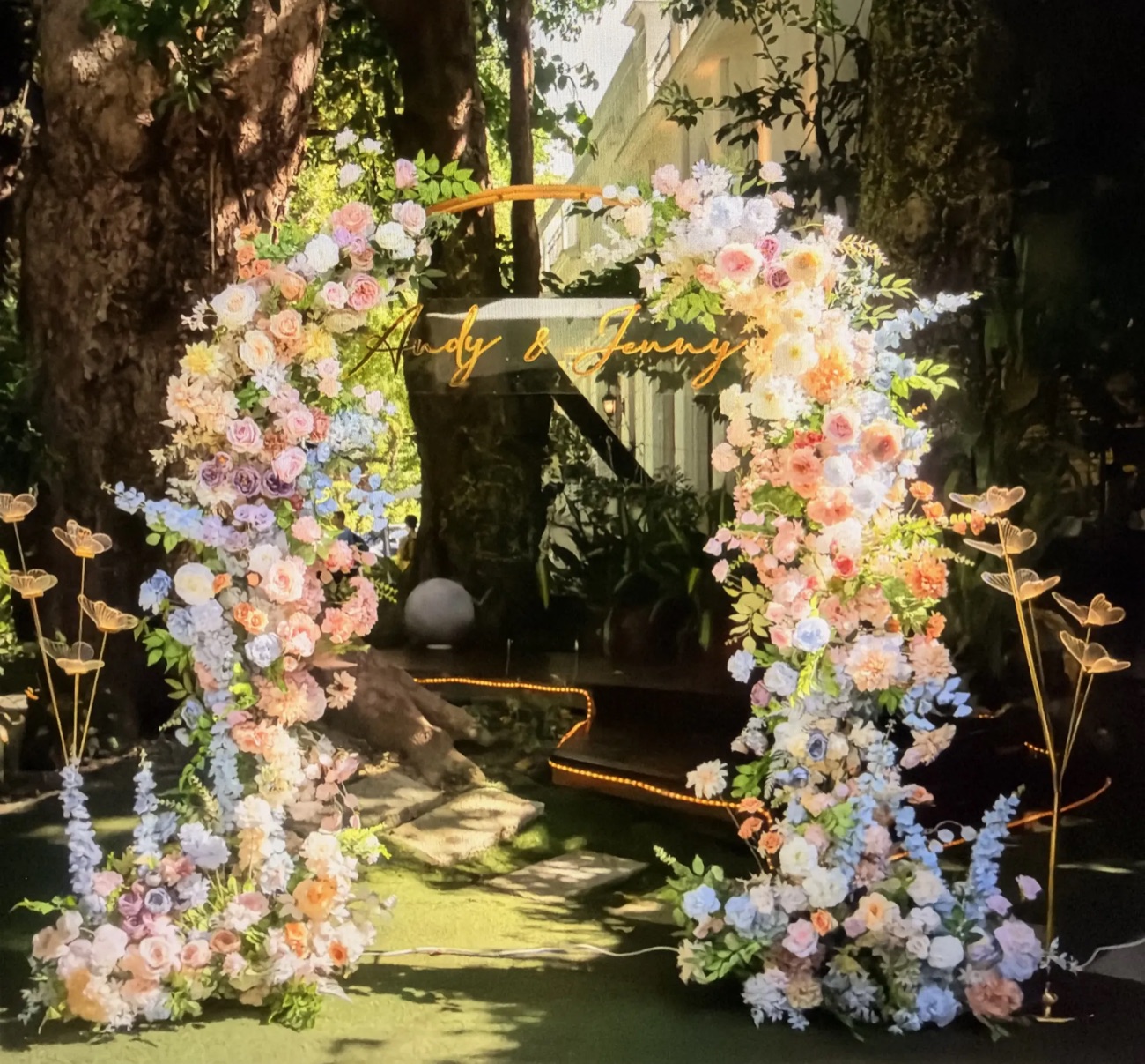 An open floral arch in blush, peach, sky blue and lavender roses and delphinium, flowing down to the grass on both sides, with gold butterfly stakes and a gold script sign