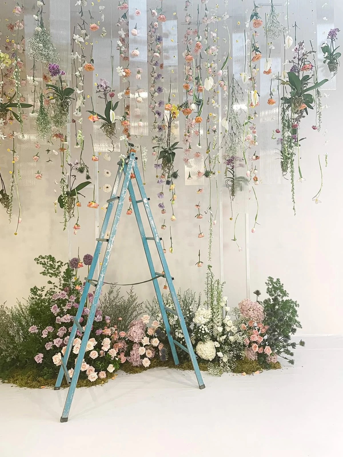Flowers in peach, lilac and white hanging on long strings, above a low bed of roses, hydrangeas and greenery on moss