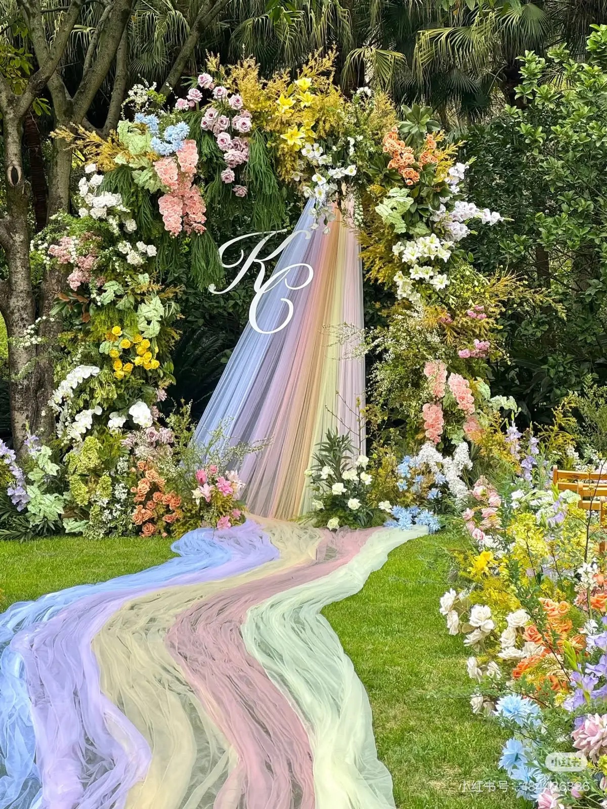 A round floral arch on a lawn in pink, peach, yellow, blue and white, with a white monogram, and pastel rainbow tulle falling from the arch and running down the aisle