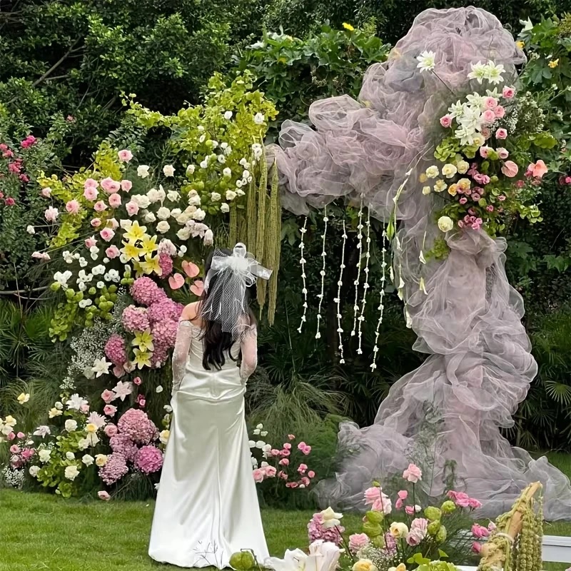 A bride in a white satin gown and veil facing an arch of lilac tulle and flowers on a lawn, with pink hydrangeas, white roses and yellow lilies on one side and strands of small white flowers hanging down