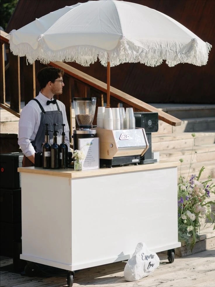 A white bar cart on wheels with a wooden top and an espresso machine, under a white umbrella with a fringed edge