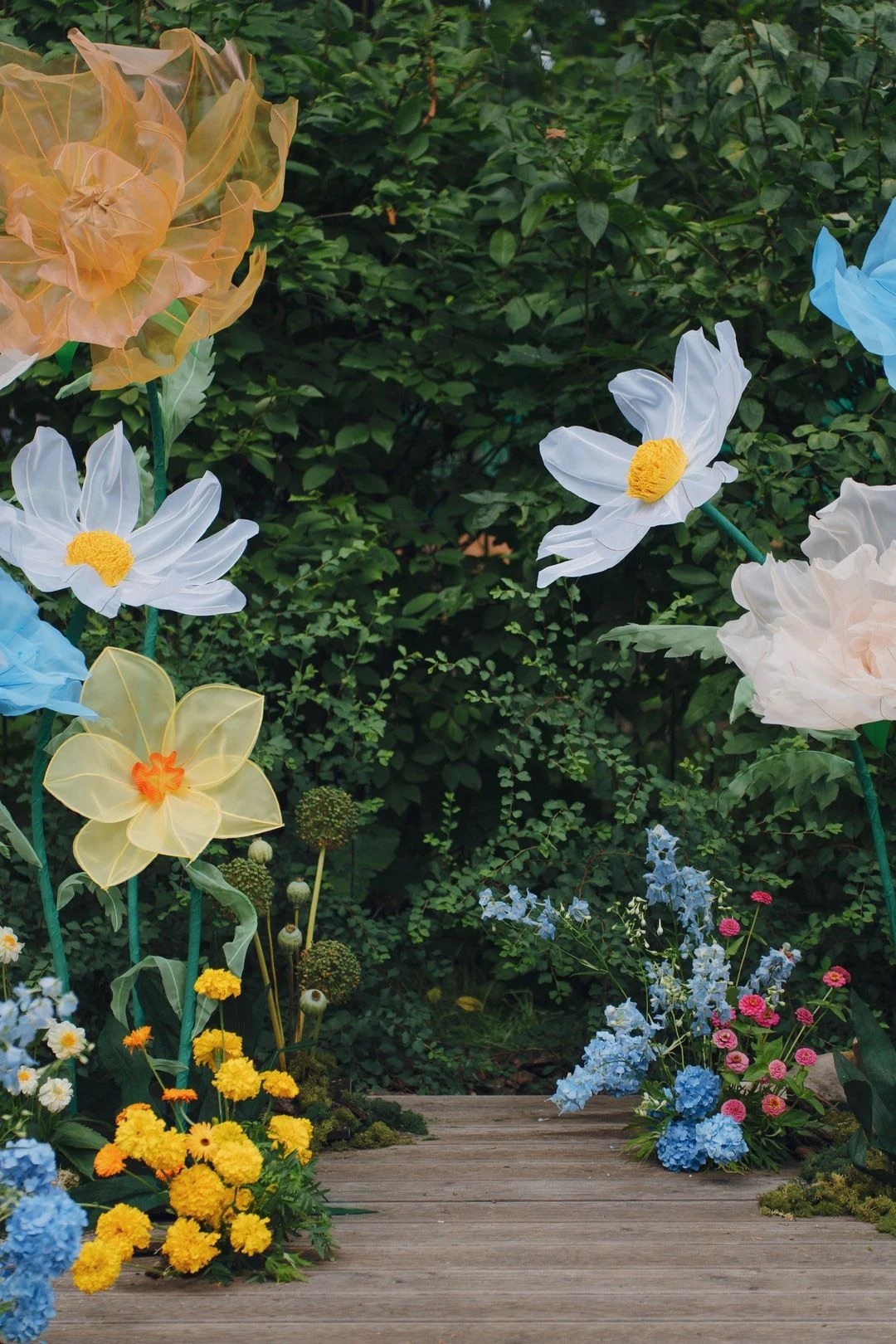 Giant sheer flowers in yellow, white, peach and blue on tall green stems, framing a wooden boardwalk edged with marigolds, hydrangeas and delphiniums