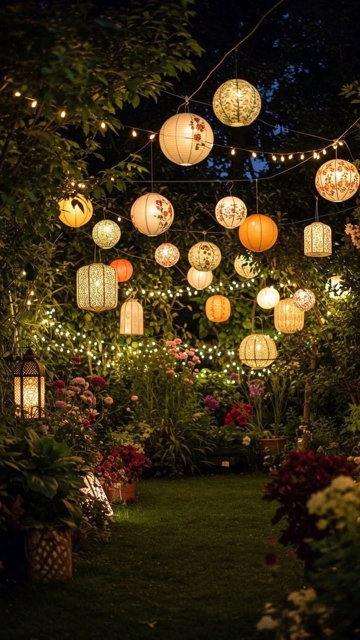 Painted paper lanterns in cream and orange hanging over a lawn at night, with string lights strung through the trees and flowers below