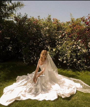 Bride seated on the grass with her dress spread out, rose hedge behind