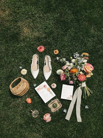 Flat lay on grass: shoes, bouquet, basket, invitation and perfume