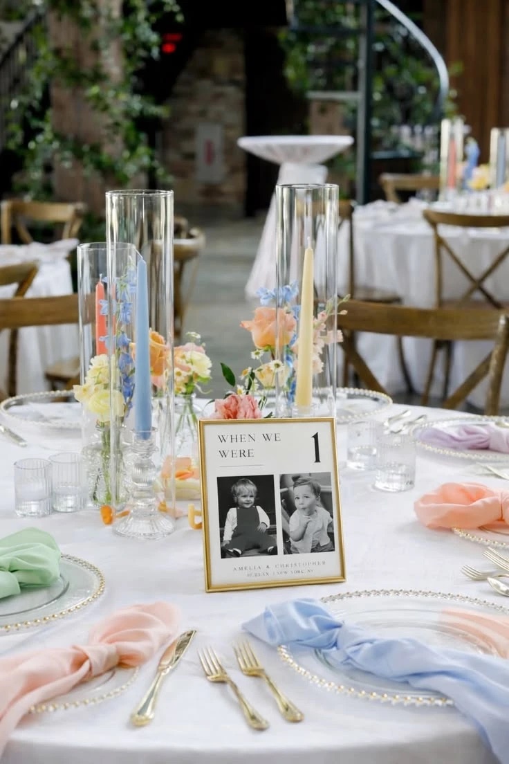 A round table in white linen with napkins in pink, blue, peach and mint, gold-rimmed glass plates, taper candles in pastel colours inside tall glass hurricanes, small vases of roses and delphiniums, and a gold-framed table number with two baby photos