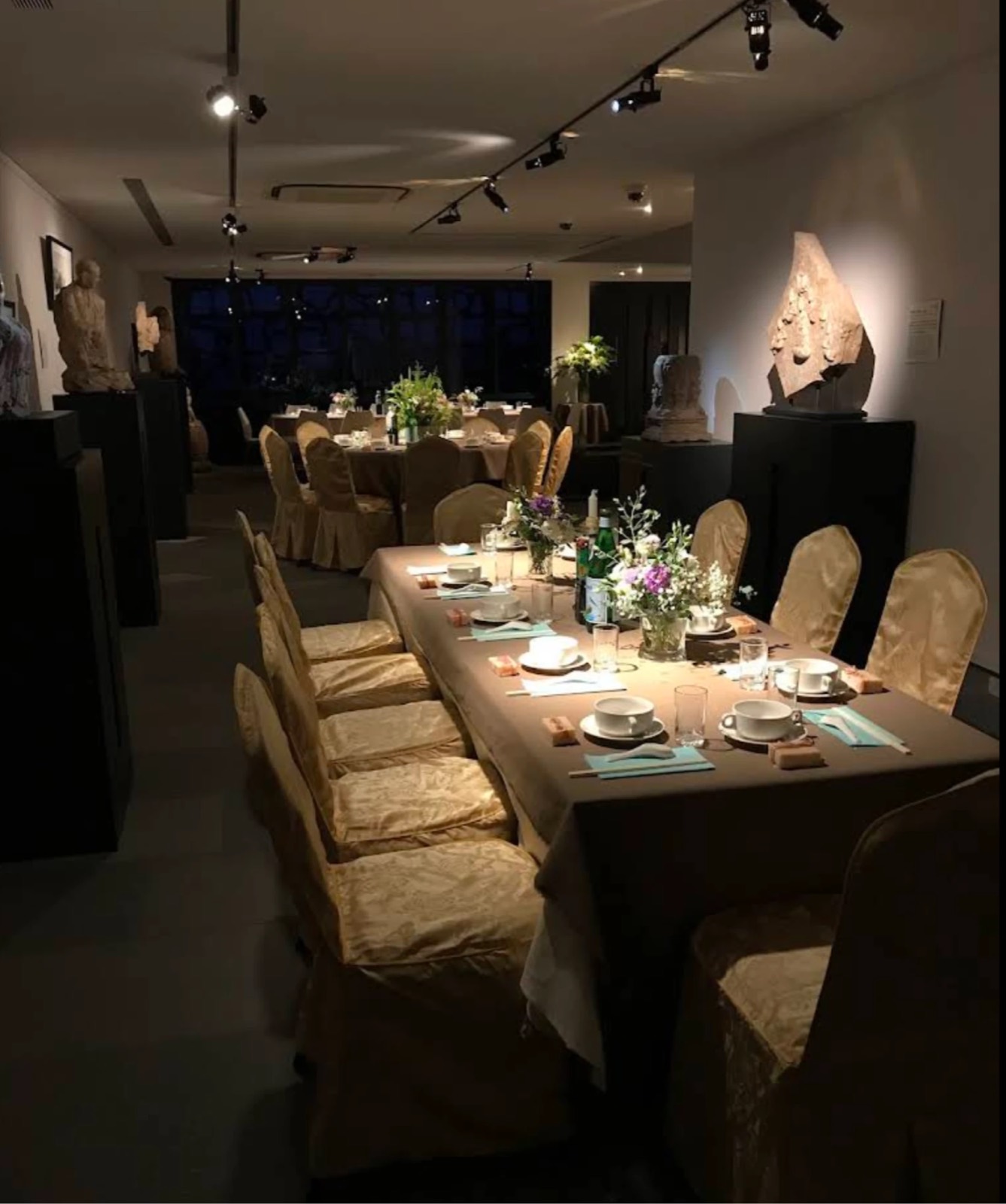 The 3rd floor set for dinner at night: long tables in taupe linen with gold chair covers, small vases of white and purple flowers, teal napkins, and spotlights on the sculptures along the walls